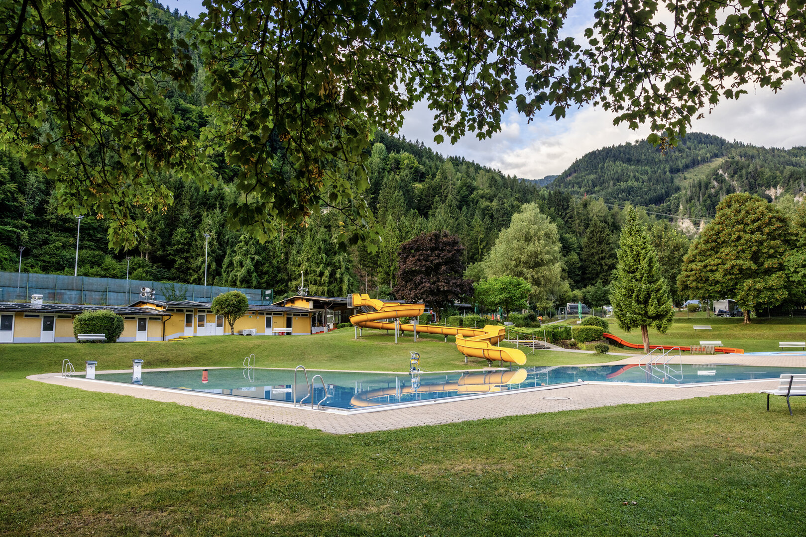 Freibad mit Wasserrutschen und Liegewiese, umgeben von Wald und Bergen
